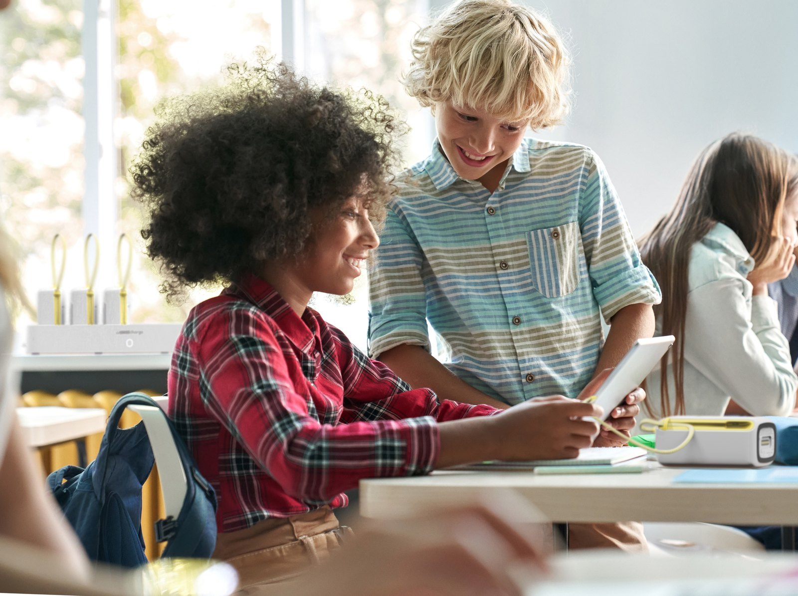 Two students sharing a tablet at a classroom desk, an Omni Locker 10E unit on the desk and a Locker Station 10E on the counter behind them
